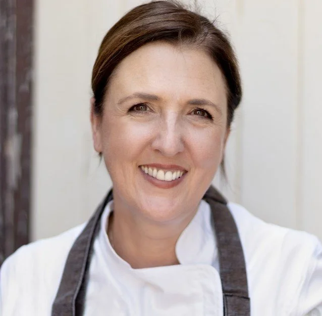 Smiling woman chef in a white chef coat and black apron outdoors, with a neutral background.