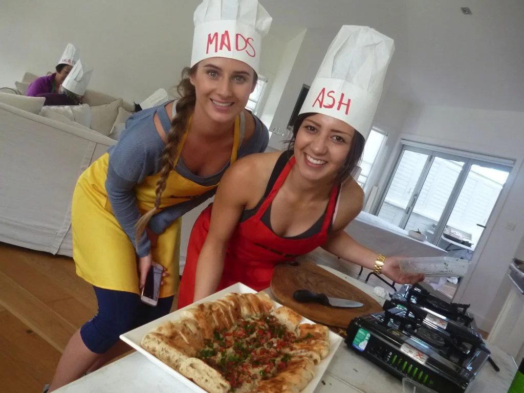 Two women wearing chef hats labeled 'MADS' and 'ASH' are smiling in a kitchen with pizza and a cutting board.