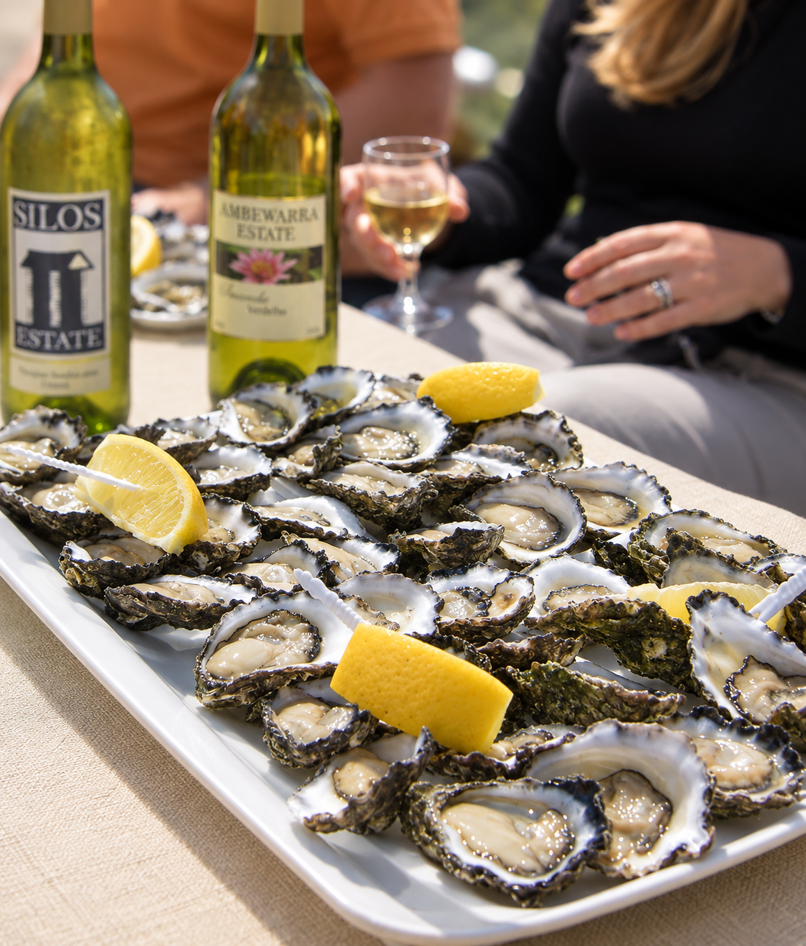 A large white platter of raw oysters on the half shell with lemon wedges, set on a beige table. In the background, two bottles of white wine, a filled wine glass, and people sitting and enjoying the meal.