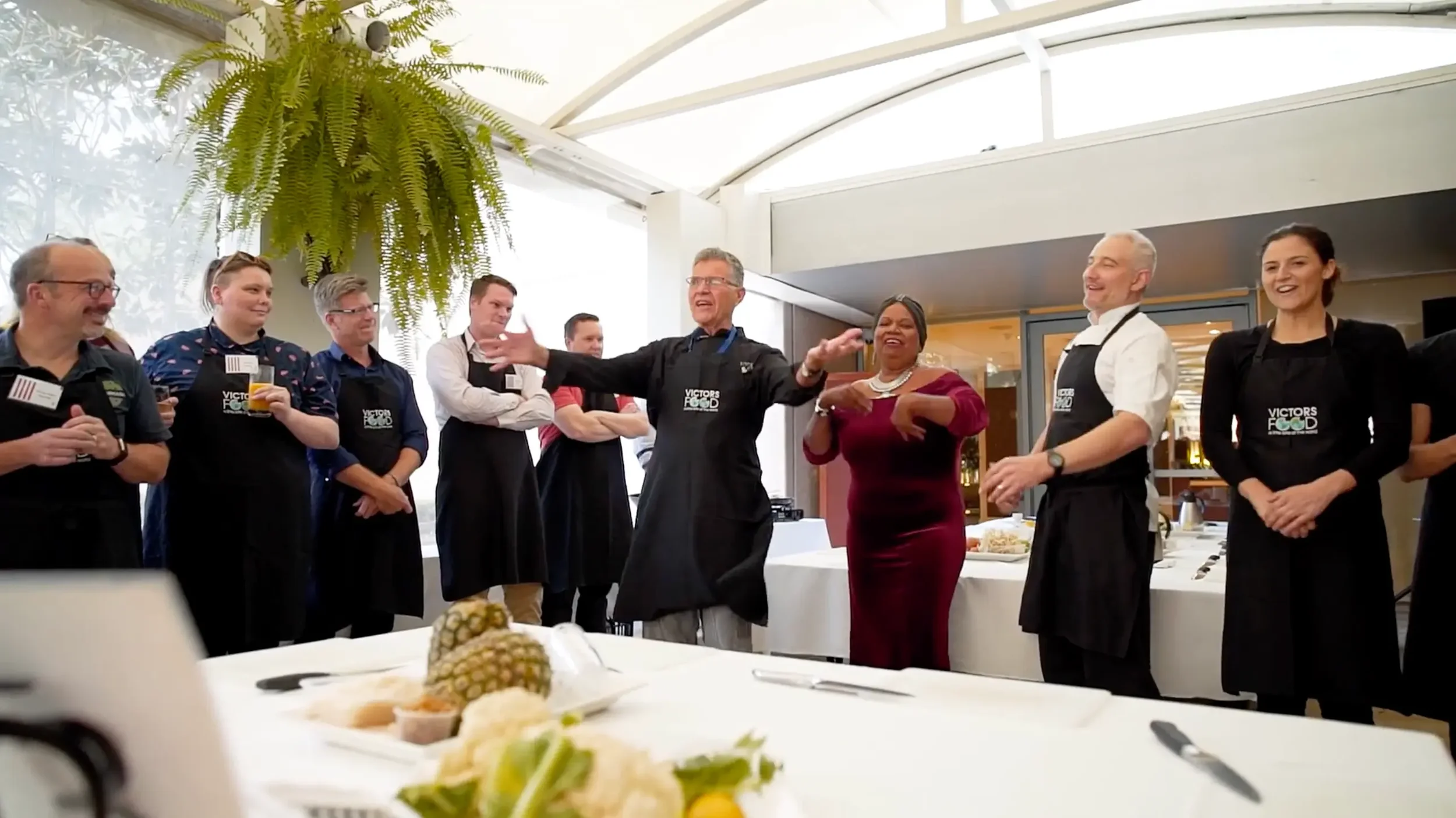 A group of people gathered in a bright indoor space, some wearing aprons, with a chef in the center speaking or presenting to the group. There are tables with food and drinks, and a large green fern hanging from the ceiling.