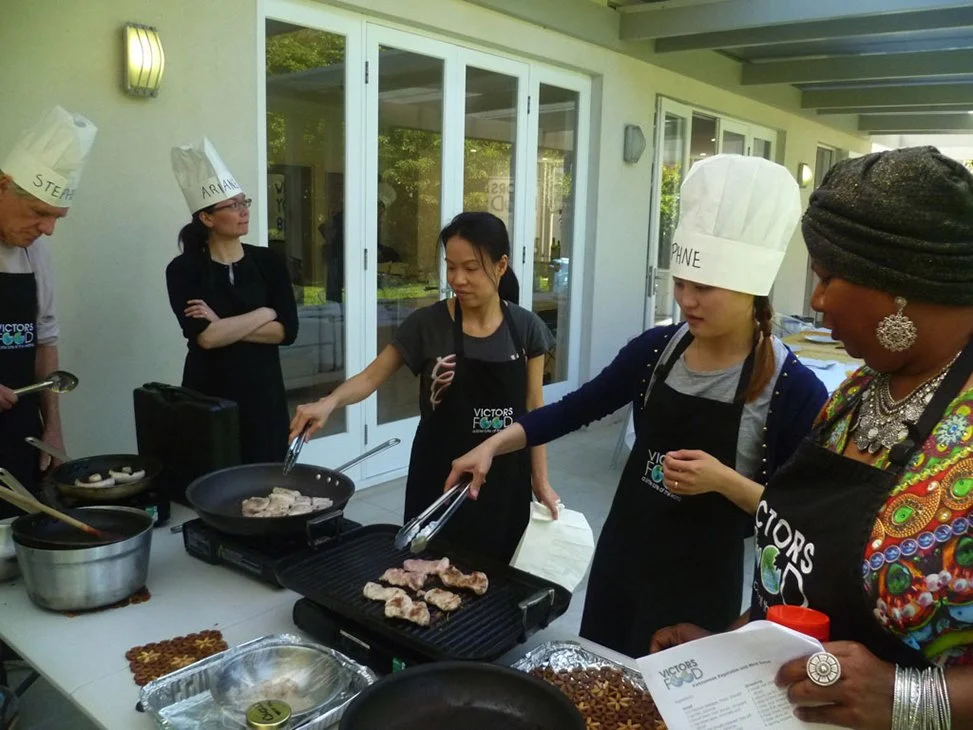 Group of people cooking outdoor, wearing chef hats and aprons, with one person flipping meat on the grill.