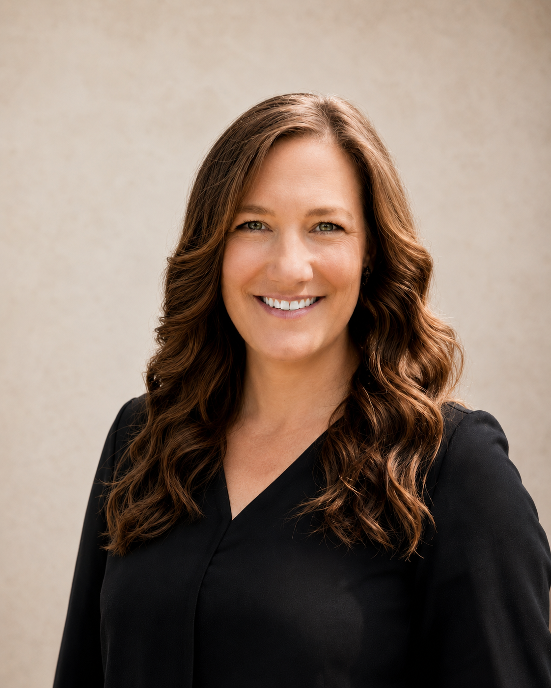 A woman with long wavy brown hair smiling, wearing a black top, standing against a plain light-colored background.