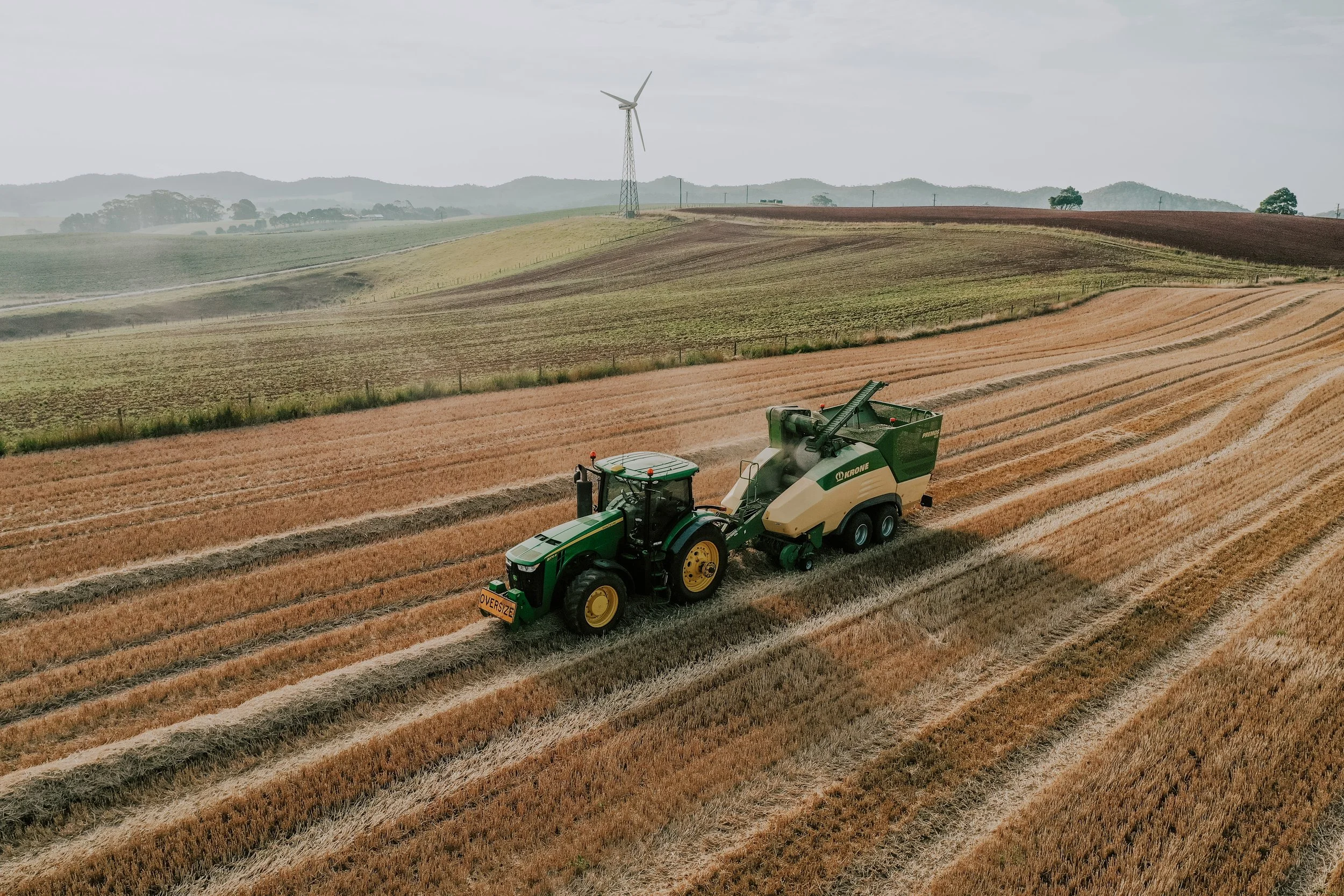 A large agricultural field with a tractor operating on it. The landscape features rolling hills, a wind turbine, and a farm vehicle working on the land. The sky is cloudy and overcast.