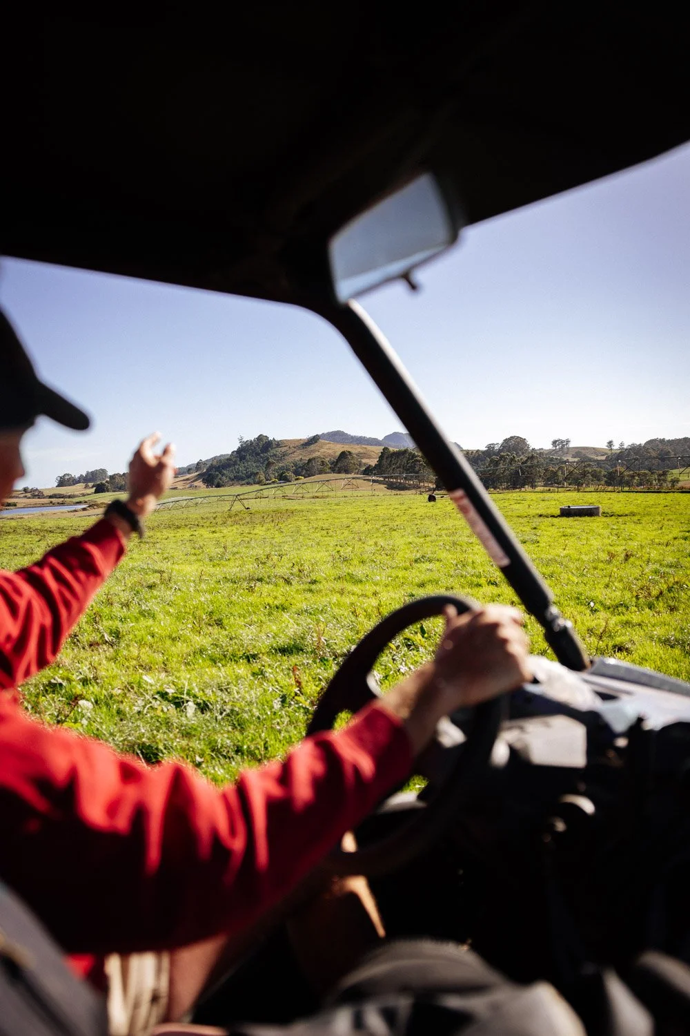 Person driving a vehicle on a grassy field, wearing a red jacket and black cap, with their hand raised and scenic hills in the background.