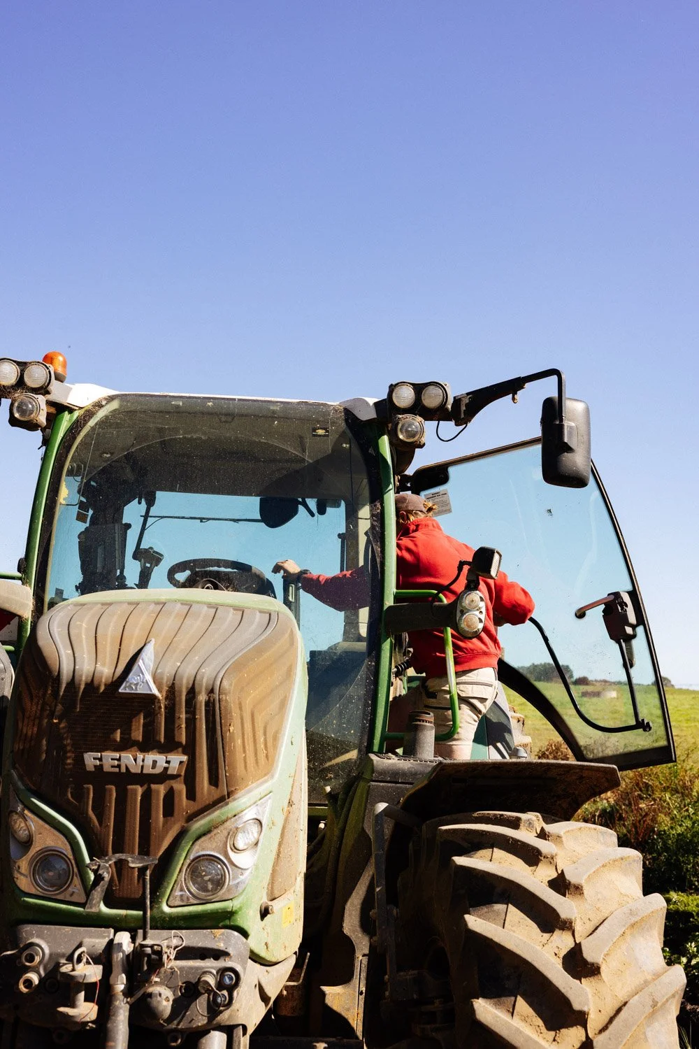A person in a red jacket and beige pants standing on a tractor, reaching inside the tractor cab, with a clear blue sky overhead.