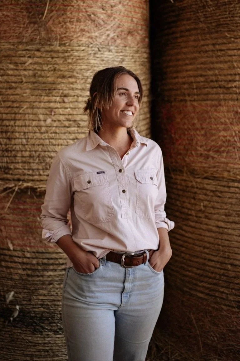 Hannah standing in a hay shed, with round bales in the background, smiling.