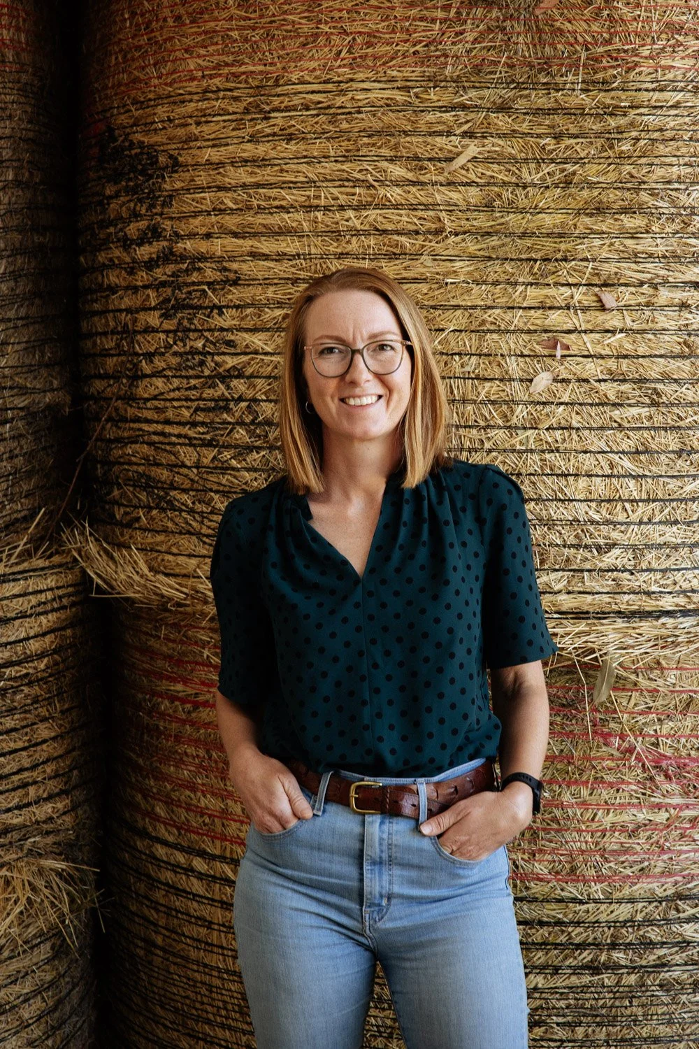 Emily standing in a hay shed, with round bales in the background, smiling.
