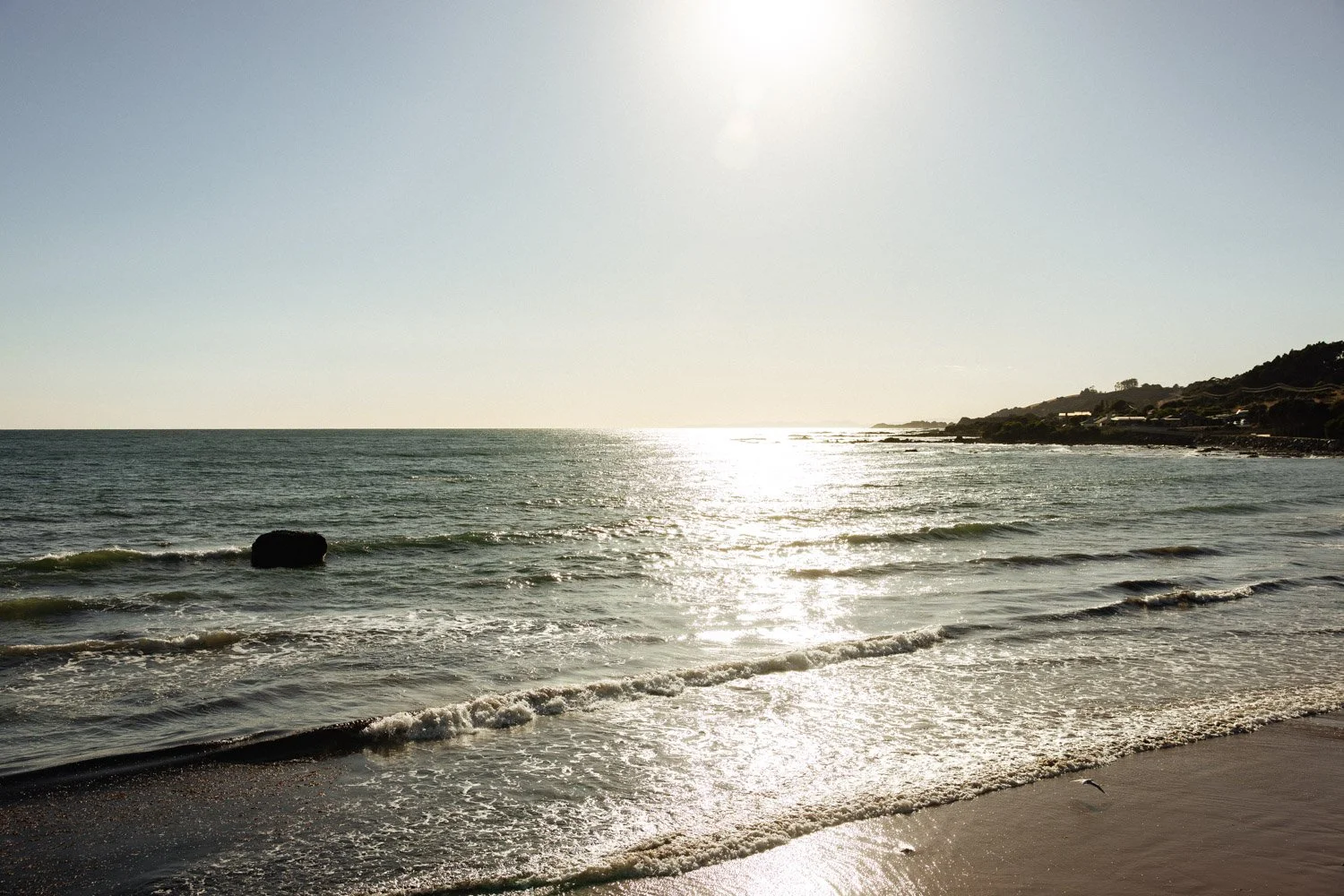 Sun reflecting on the ocean near a sandy beach with small waves and a rock in the water, with hills and houses in the background under a clear sky.