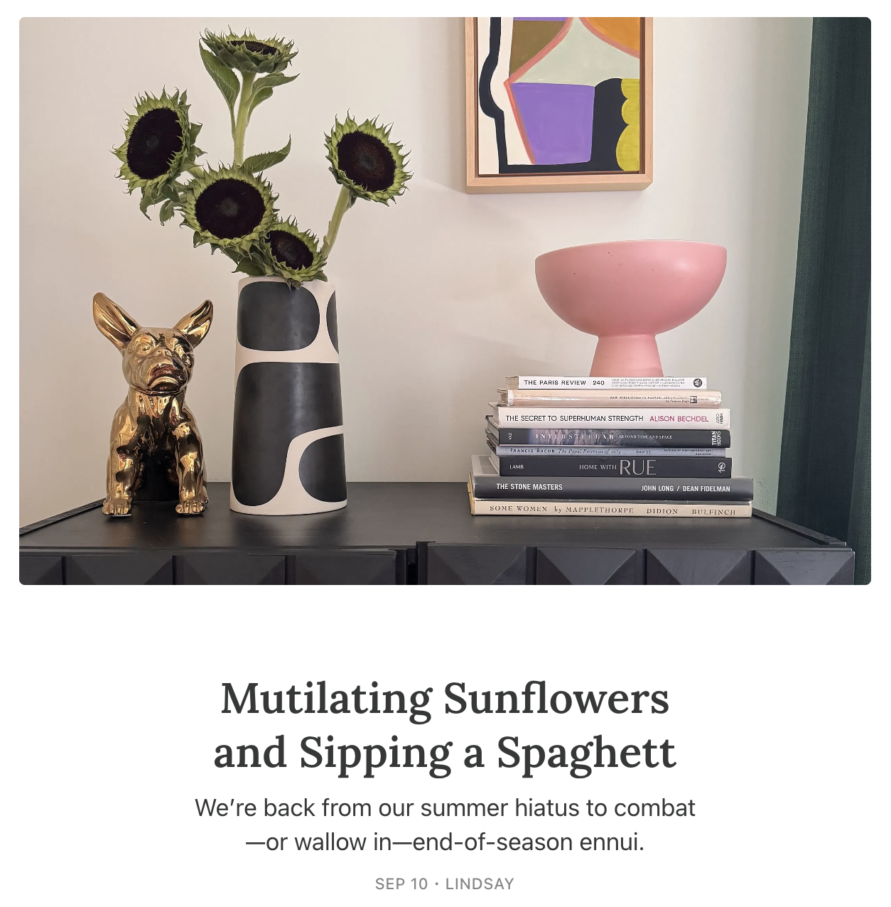 Decorative tabletop with a black sideboard, featuring a black and white vase with sunflowers, a gold French bulldog figurine, a pink bowl, and a stack of books. In the background, there is an abstract colorful painting on the wall and green curtains.