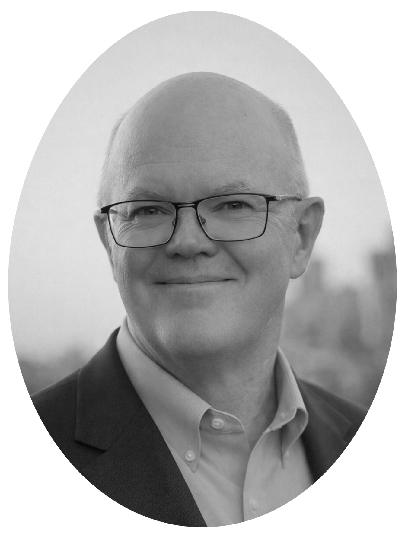 Black and white professional headshot of a bald man wearing glasses, a suit, and a dress shirt, smiling slightly, outdoors with a blurred cityscape background.