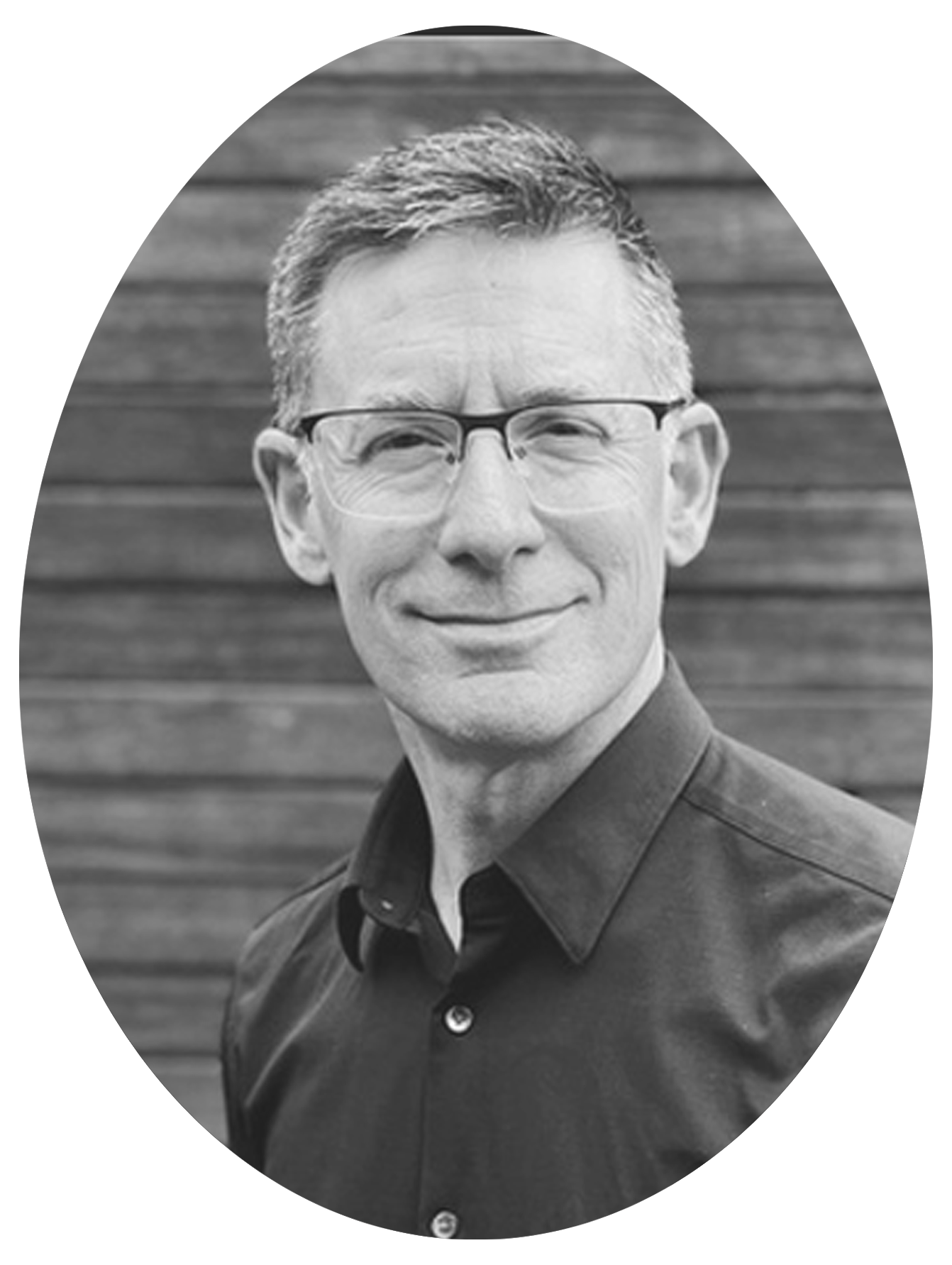 Black and white portrait of a middle-aged man with short, tousled hair, wearing glasses, smiling, with a wooden background.