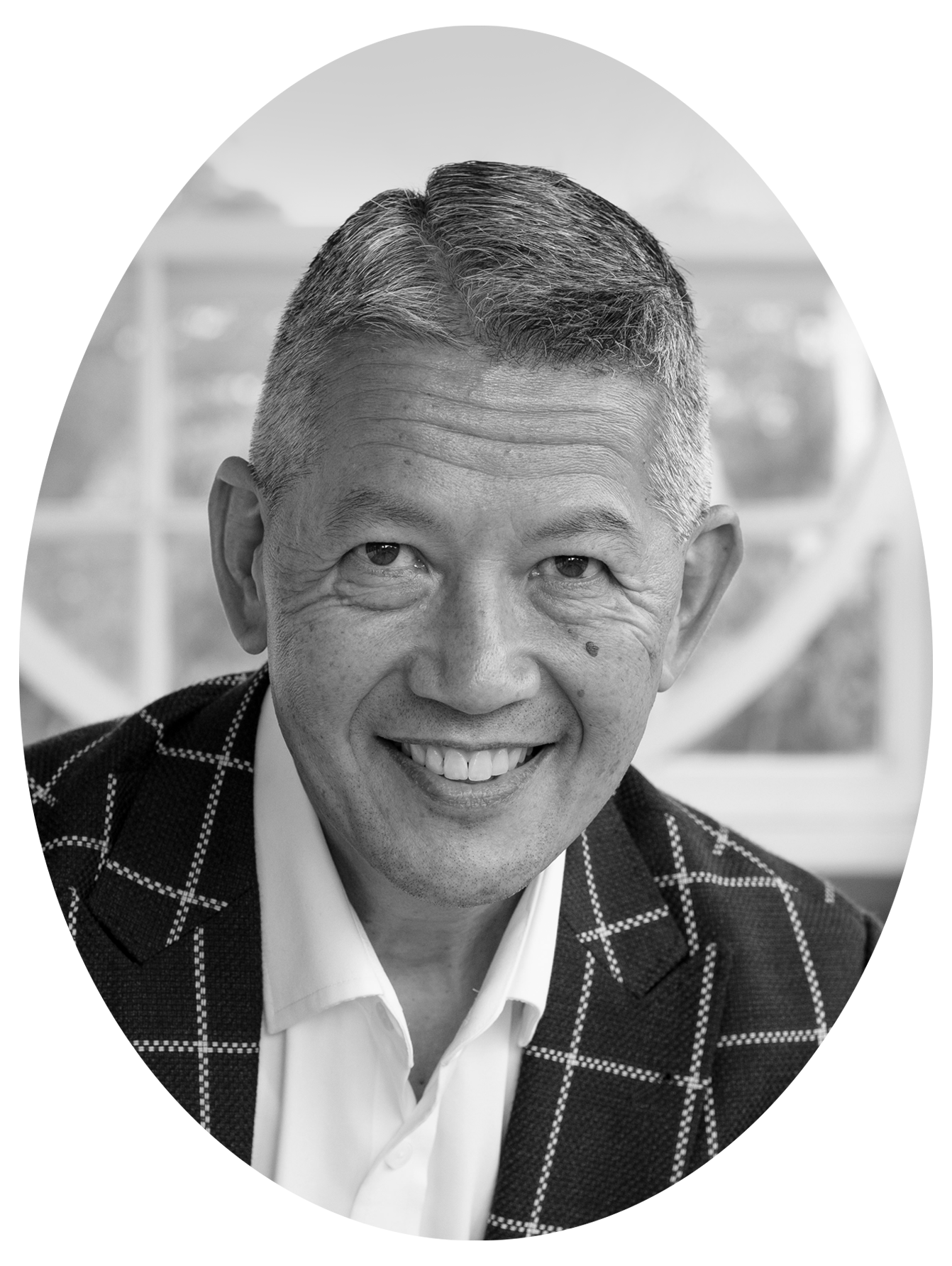 Black and white portrait of a smiling man with short hair, wearing a checkered blazer and a white shirt, in front of a blurred background.