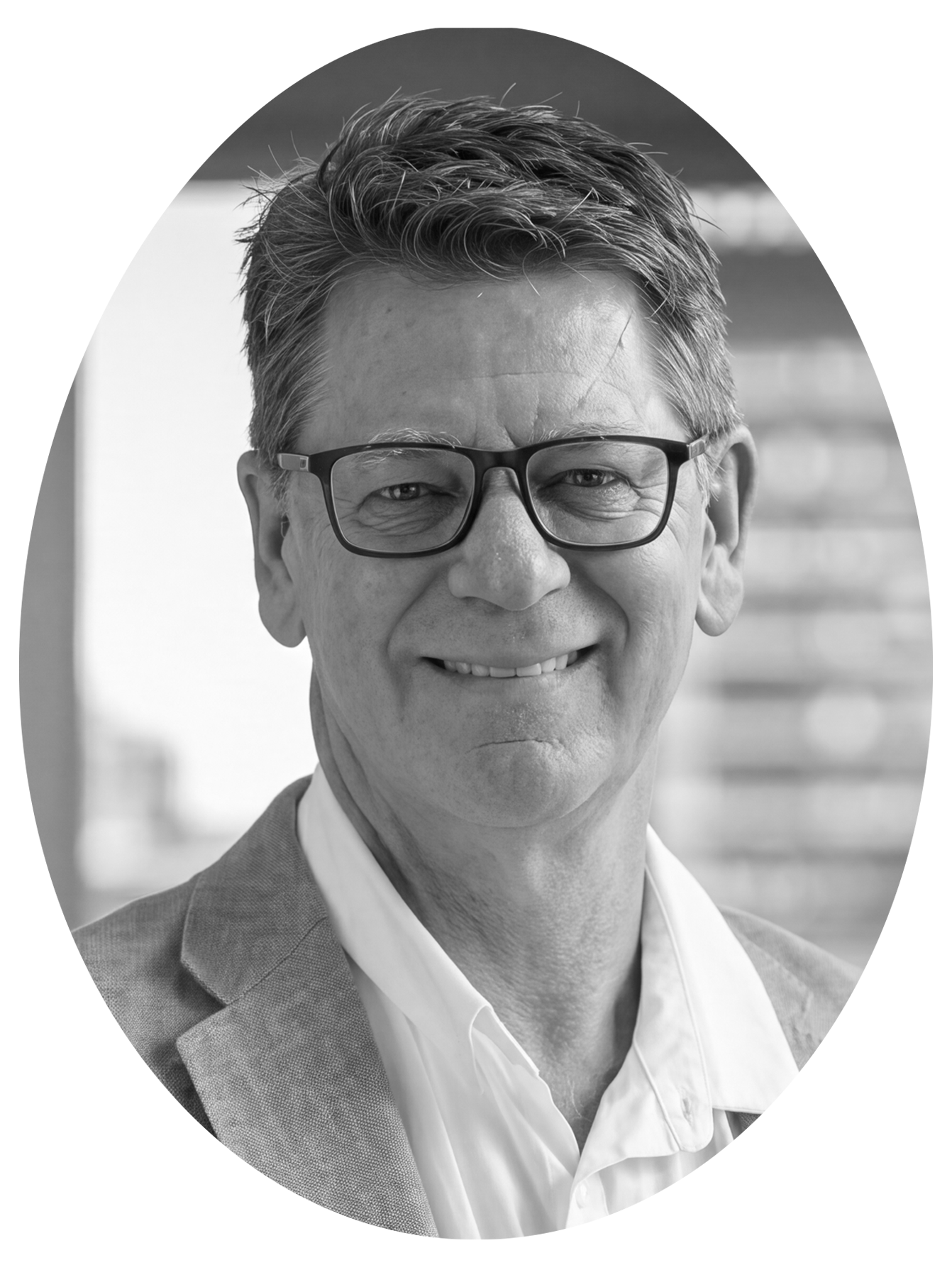 Black and white portrait of a middle-aged man with glasses, smiling, wearing a suit jacket and white shirt, standing outdoors with blurred background.