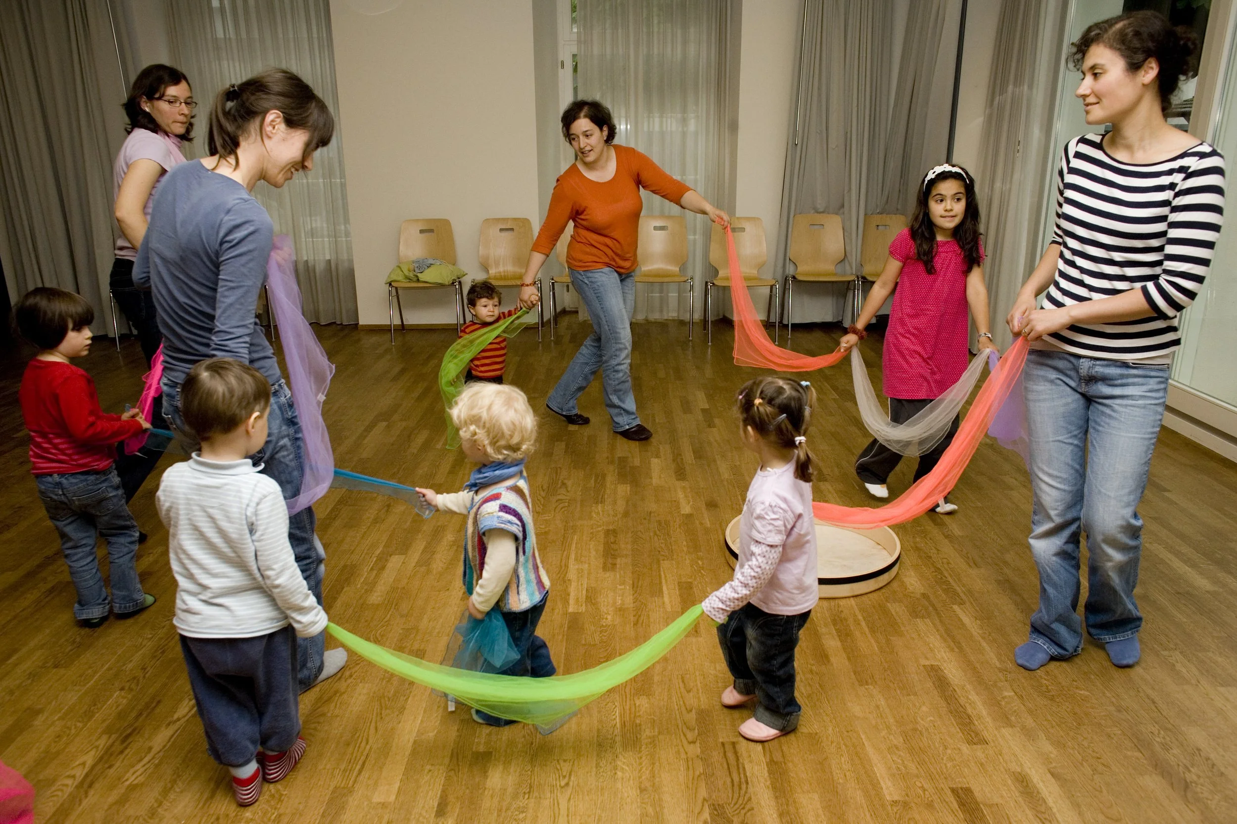 Kinder und Erwachsene spielen in einem Raum mit bunten Tüchern und einer runden Plattform, halten sich an die Tücher und umkreisen die Plattform.