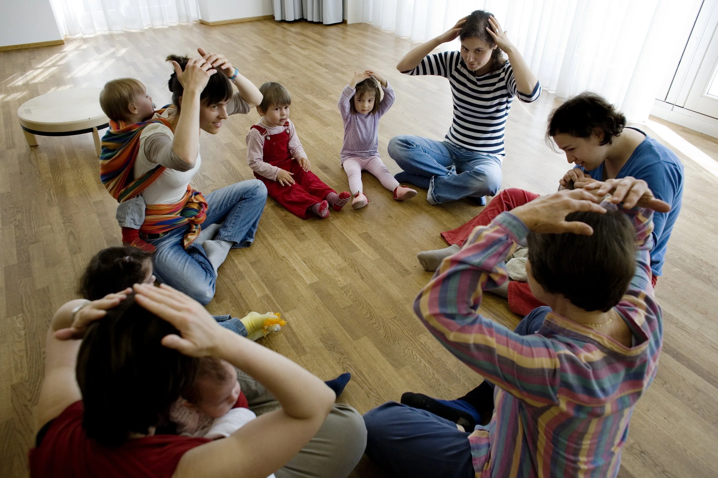 Eine Gruppe von Erwachsenen und Kindern sitzen im Kreis auf dem Boden in einem Raum mit Holzboden und weißen Vorhängen. Die Erwachsenen scheinen eine Aktivität anzuleiten, während die Kinder aufmerksam zuhören.
