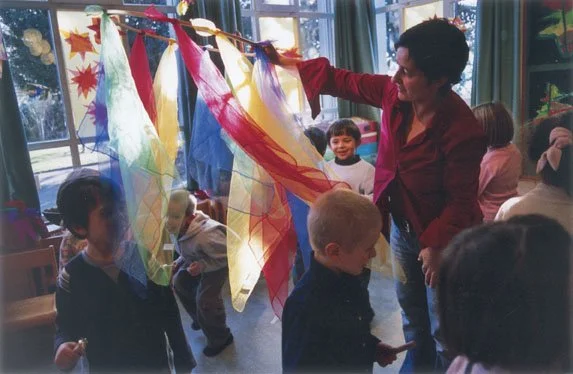 Eine Frau zeigt bunten, transparenten Stoff oder Papier an einer Schnur in einem Raum mit Kindern, die zuschauen.