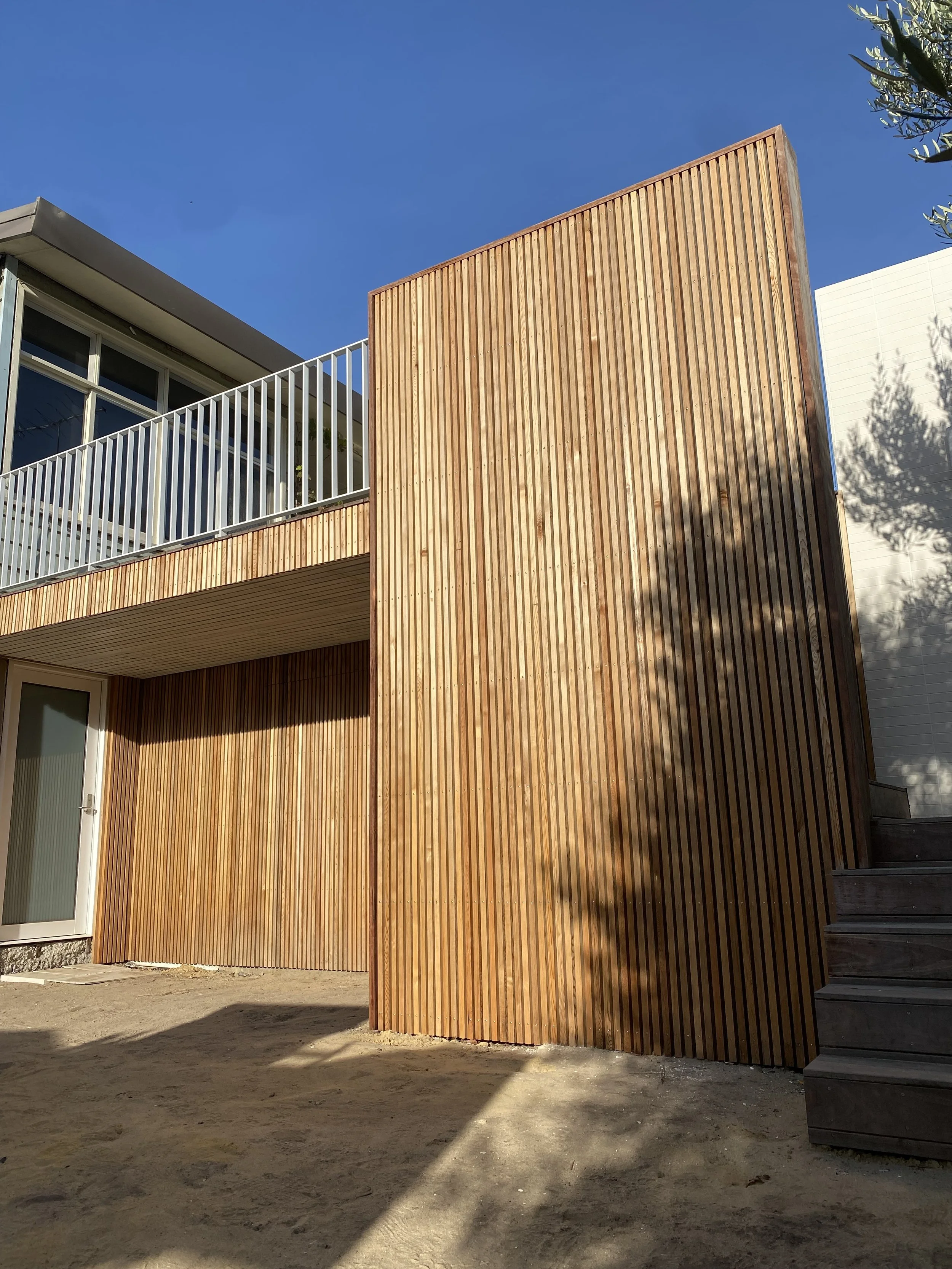 Modern building with wooden vertical slats on exterior wall, white balcony railing, glass door and window, stairs on the right, under a clear blue sky, shadows cast on ground.