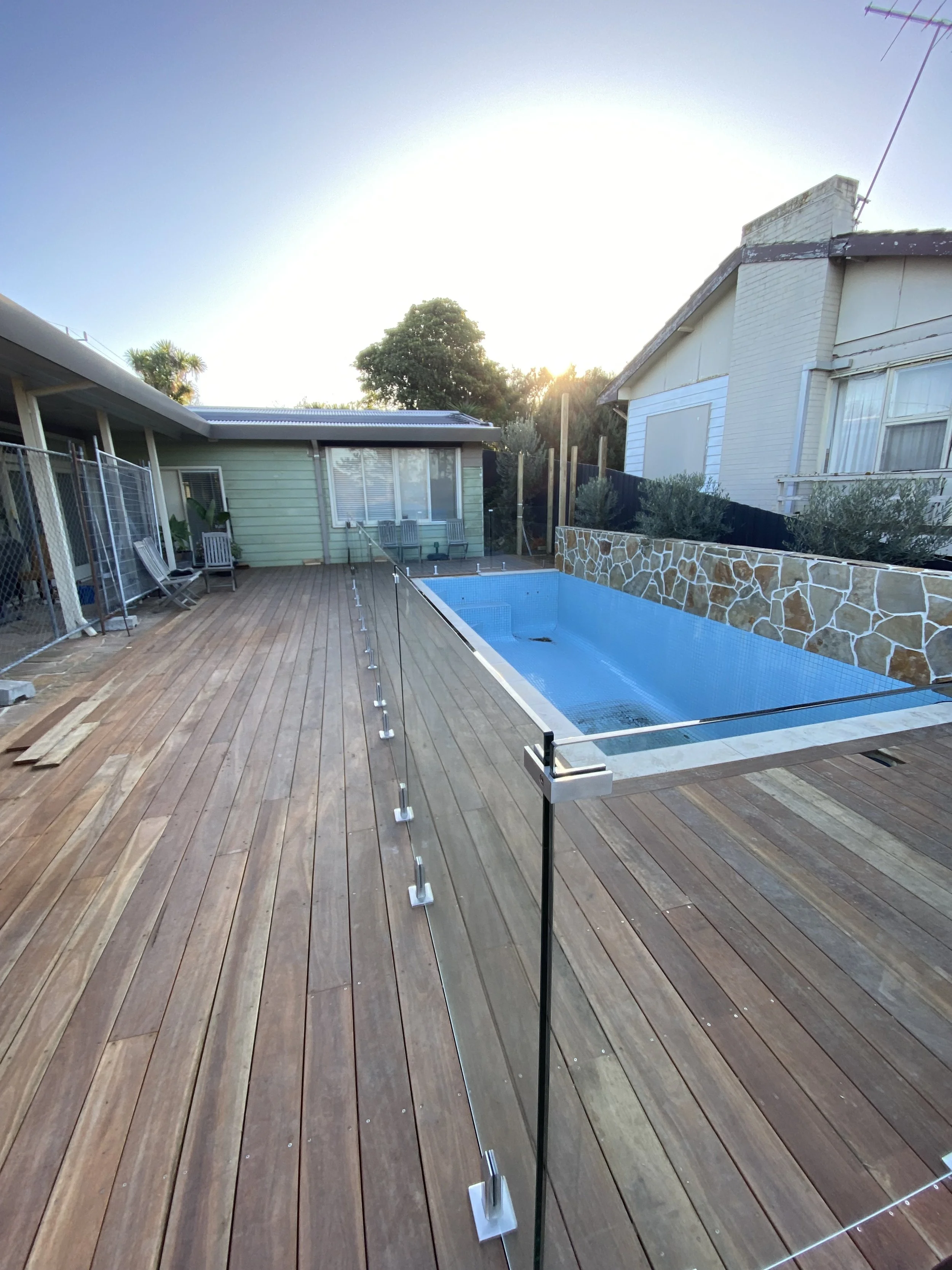 Backyard with a wooden deck, small pool, house with white and green exterior, and a clear sky with the sun setting or rising.