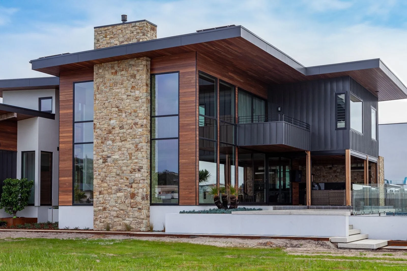 Modern house with stone, wood, and dark metal siding, large windows, small balcony, and landscaped yard.