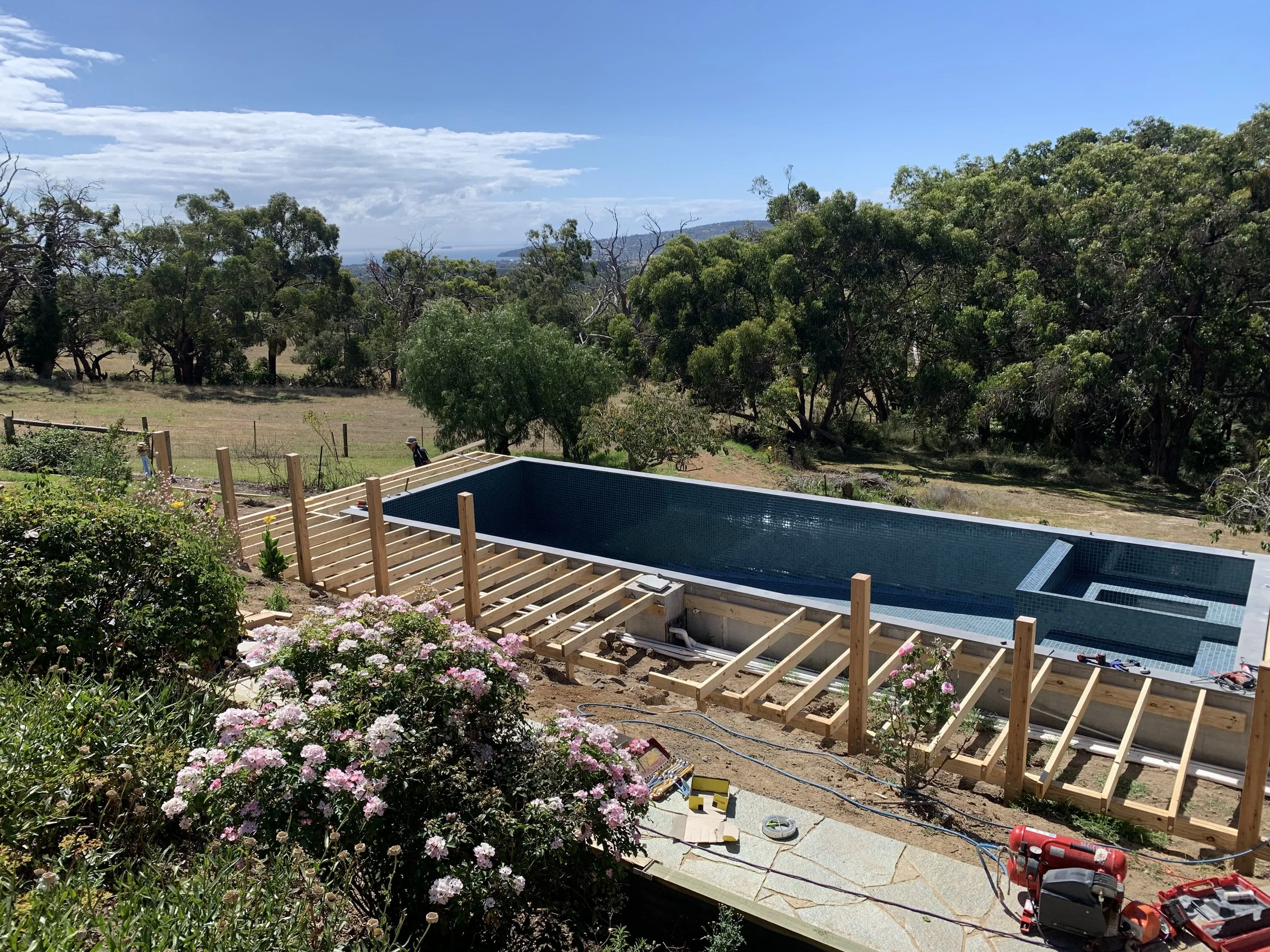 Backyard swimming pool under construction surrounded by trees and flowering plants, with tools and materials laying on the ground.