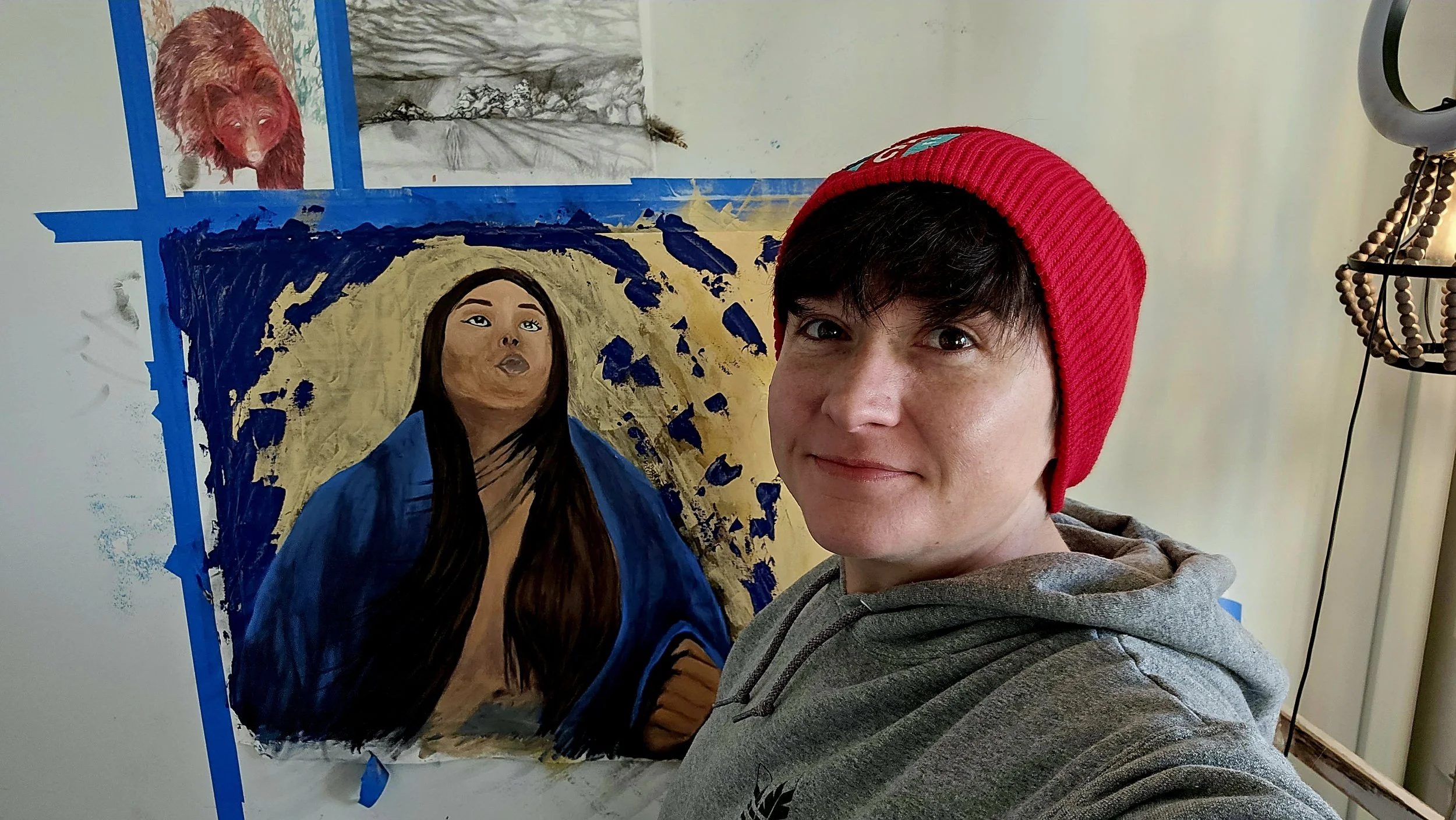 A person with short dark hair, wearing a red beanie and gray hoodie, is taking a selfie in front of two artworks on a wall. One artwork is a realistic portrait of a woman with long dark hair, and the other appears to be a nature scene. There is also a small picture of a bear in the upper left corner.