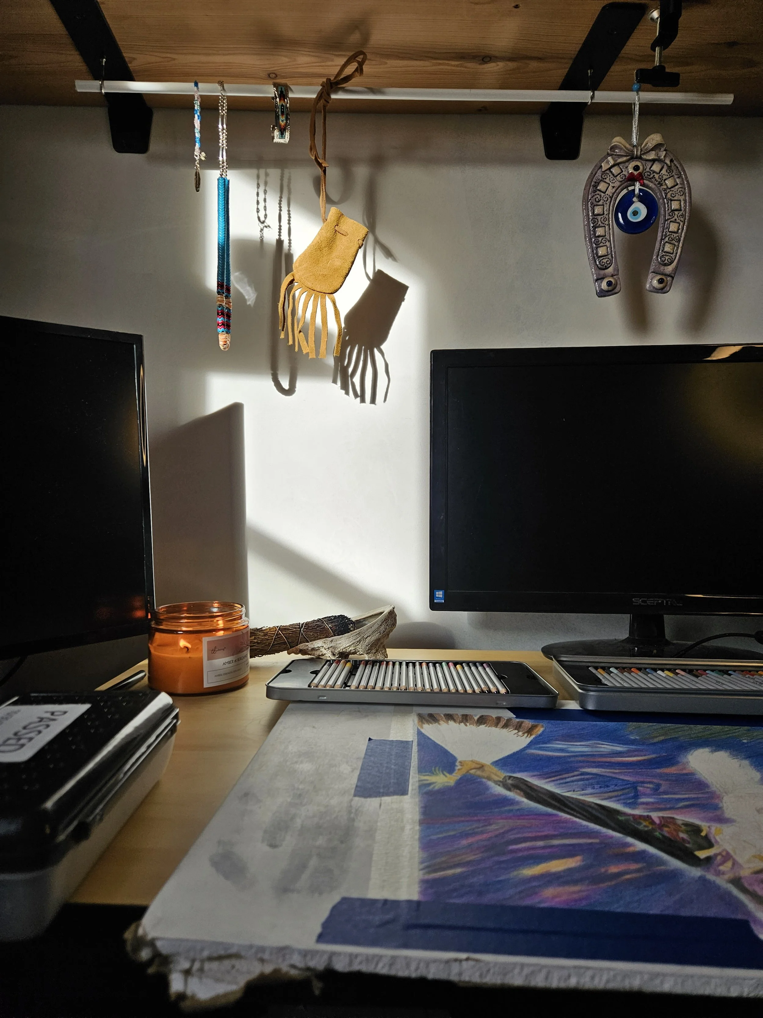 A desk with two computer monitors, a box of colored pencils, a candle, and a colorful print or painting partially visible. Hanging above are jewelry items and a hanging decorative horseshoe with an eye charm.