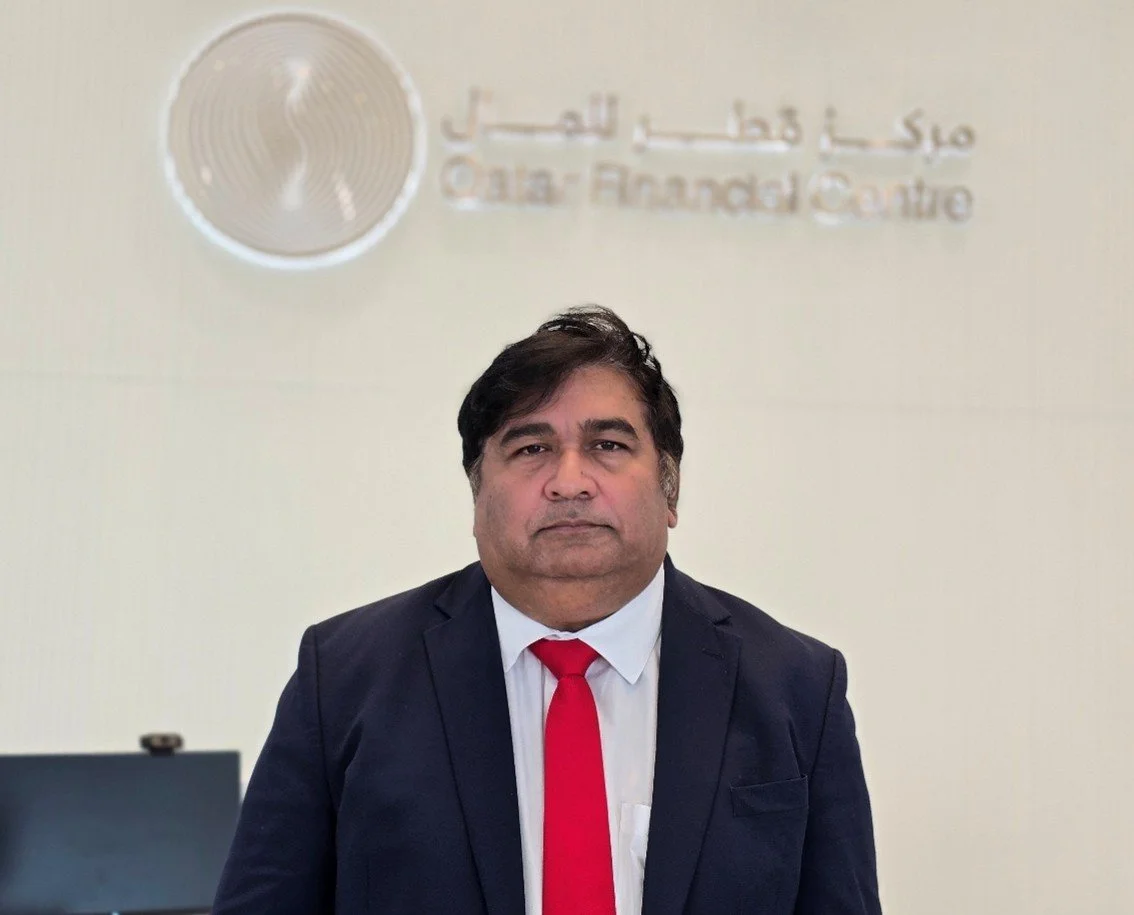 A man in a navy blue suit and red tie standing in front of a beige wall with a logo and the words 'Jamal Jhas in the Sud Air-Frouad Centro' on it.