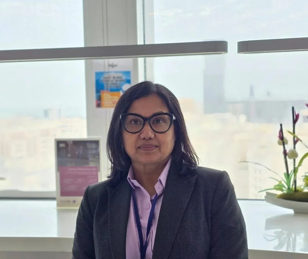 A woman with shoulder-length dark hair and glasses wearing a black blazer and light purple shirt, sitting in front of a window with city buildings outside, and a potted orchid on her right.