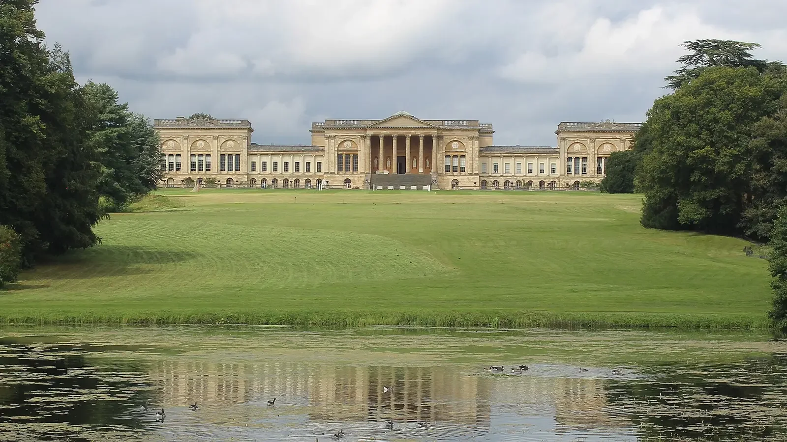 A large, historic school with classical architecture, surrounded by a green lawn, trees, and a pond with ducks in the foreground.