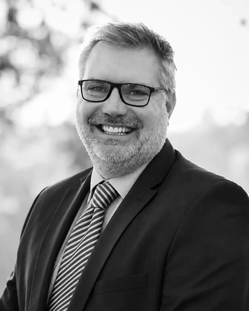 Black and white portrait of James Pym, School Expert, a suit, and a striped tie, with a blurred outdoor background.