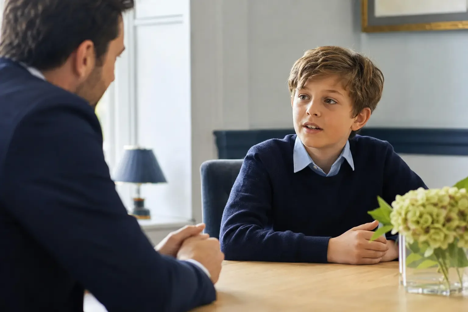 A young boy talking with an adult man across a table in a bright room, with a flower arrangement in the foreground.