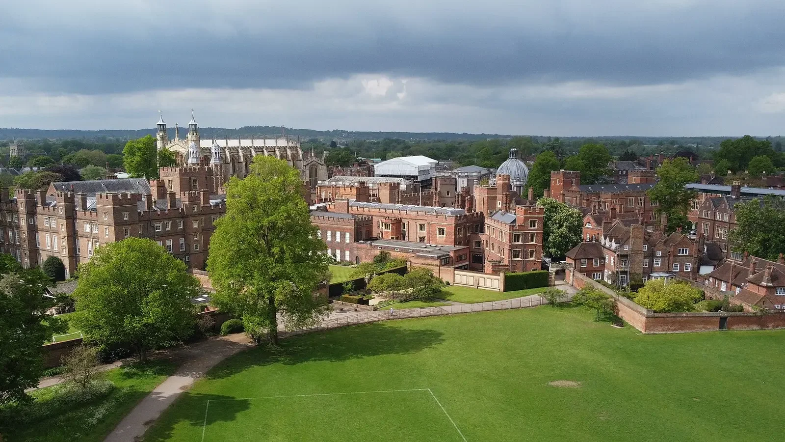 Aerial view of historic school, green trees, and a grassy park, with a cathedral and cloudy sky in the background.