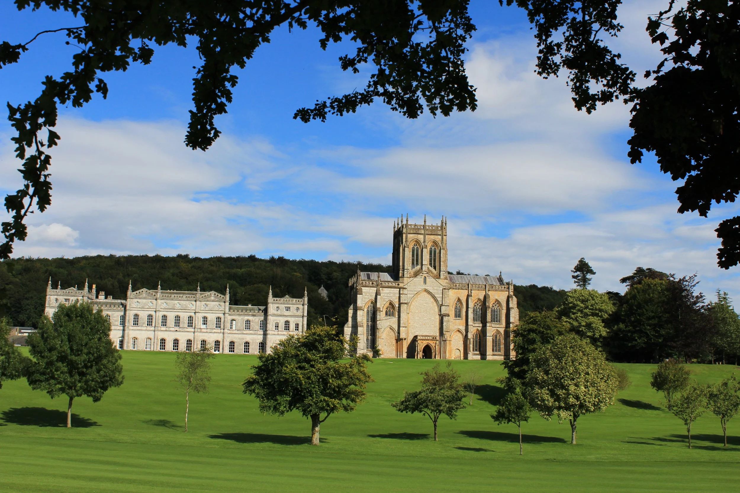 A historic school with Gothic architectural features, situated in a landscaped green area with trees, under a partly cloudy sky.