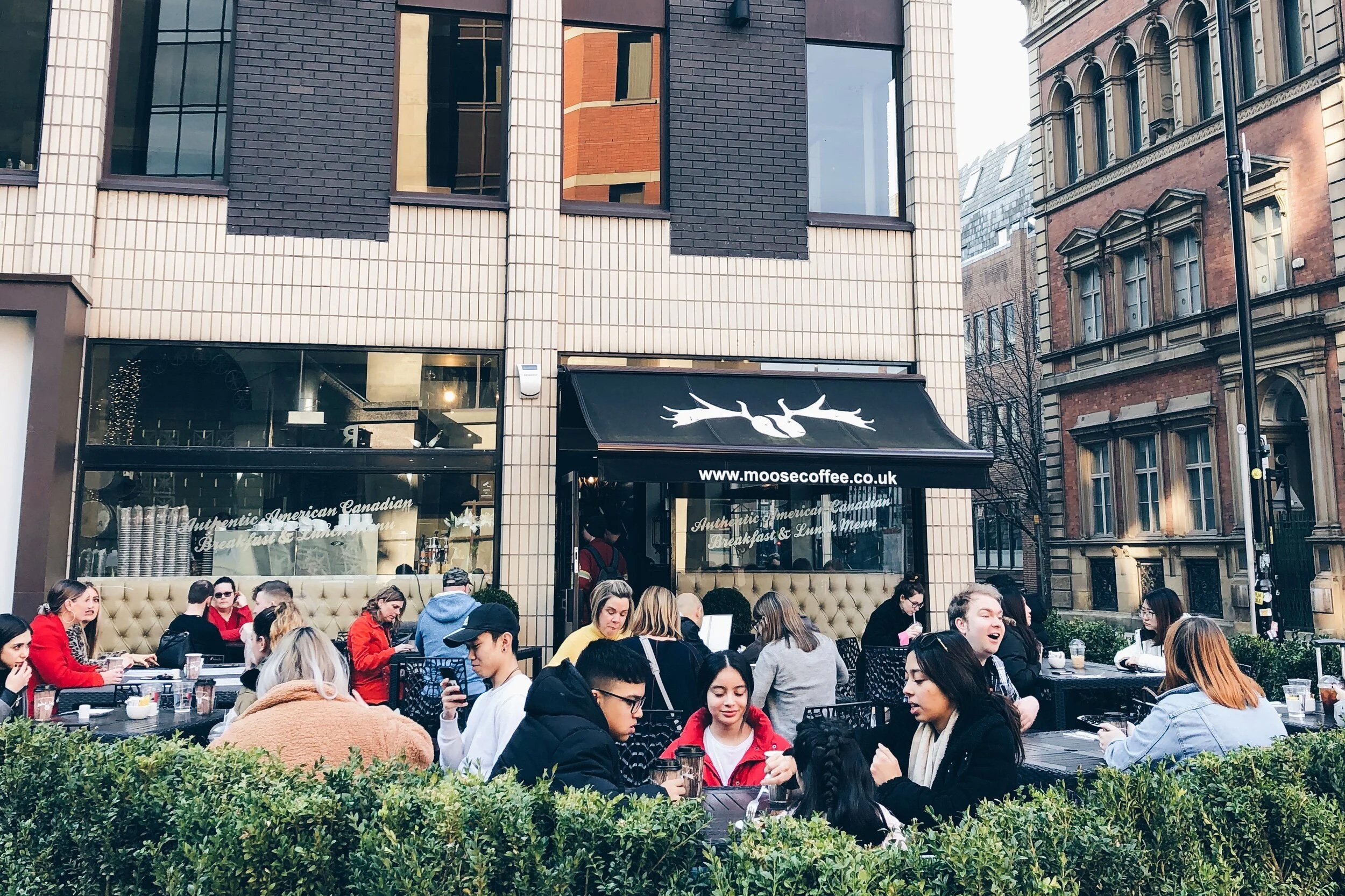 Outdoor café scene with many people seated at tables, some are talking, some are looking at their phones. The café has a black awning with a white moose logo and the website www.moosecoffee.co.uk. The background shows multi-story buildings with large windows and a modern black and white facade.