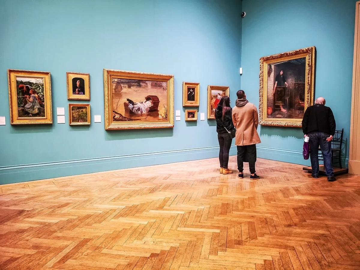 People viewing paintings in an art gallery with a wooden floor and teal walls.