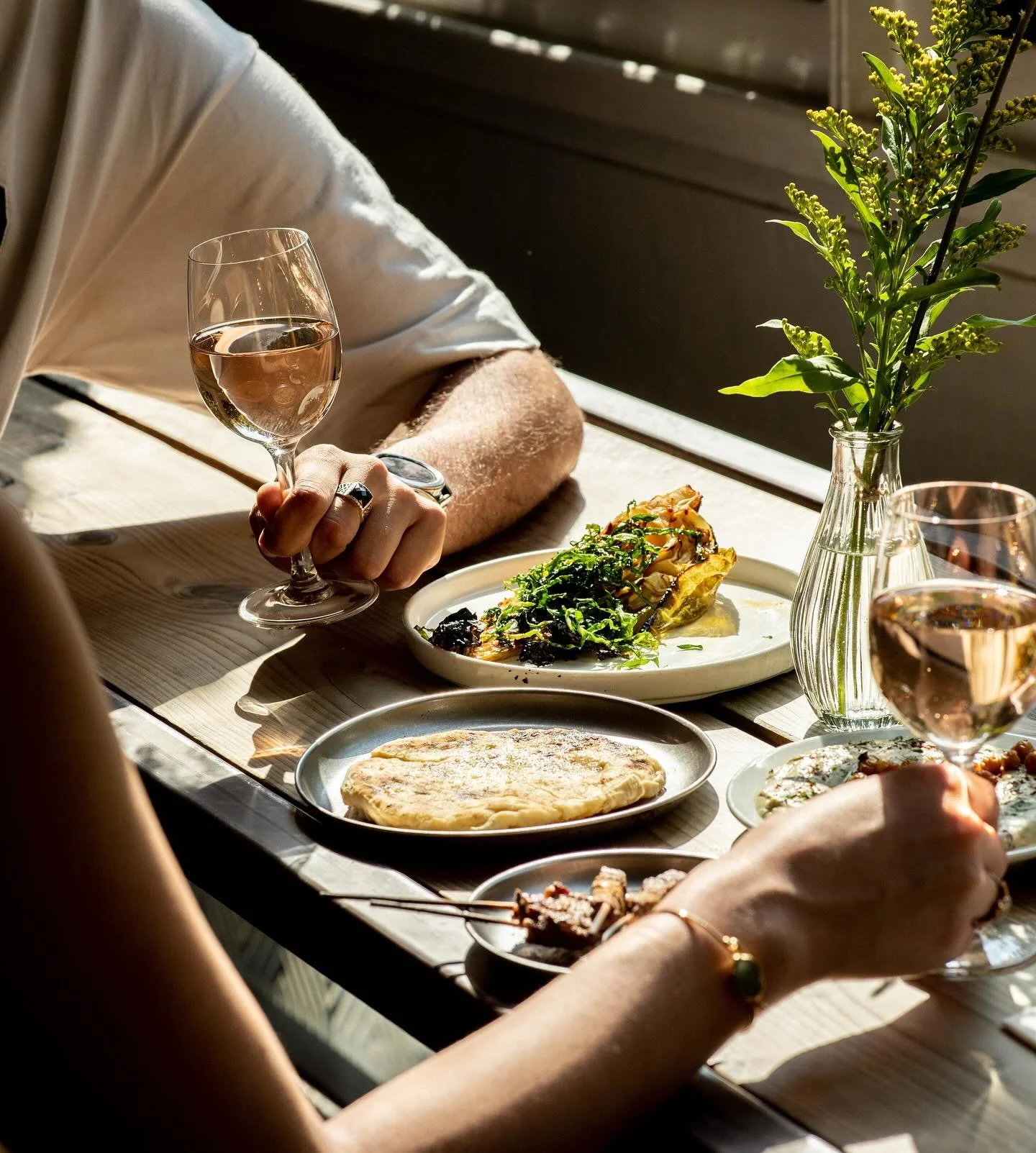 People are enjoying a meal outdoors with plates of food, glasses of rosé wine, and a small vase with greenery on a wooden table.