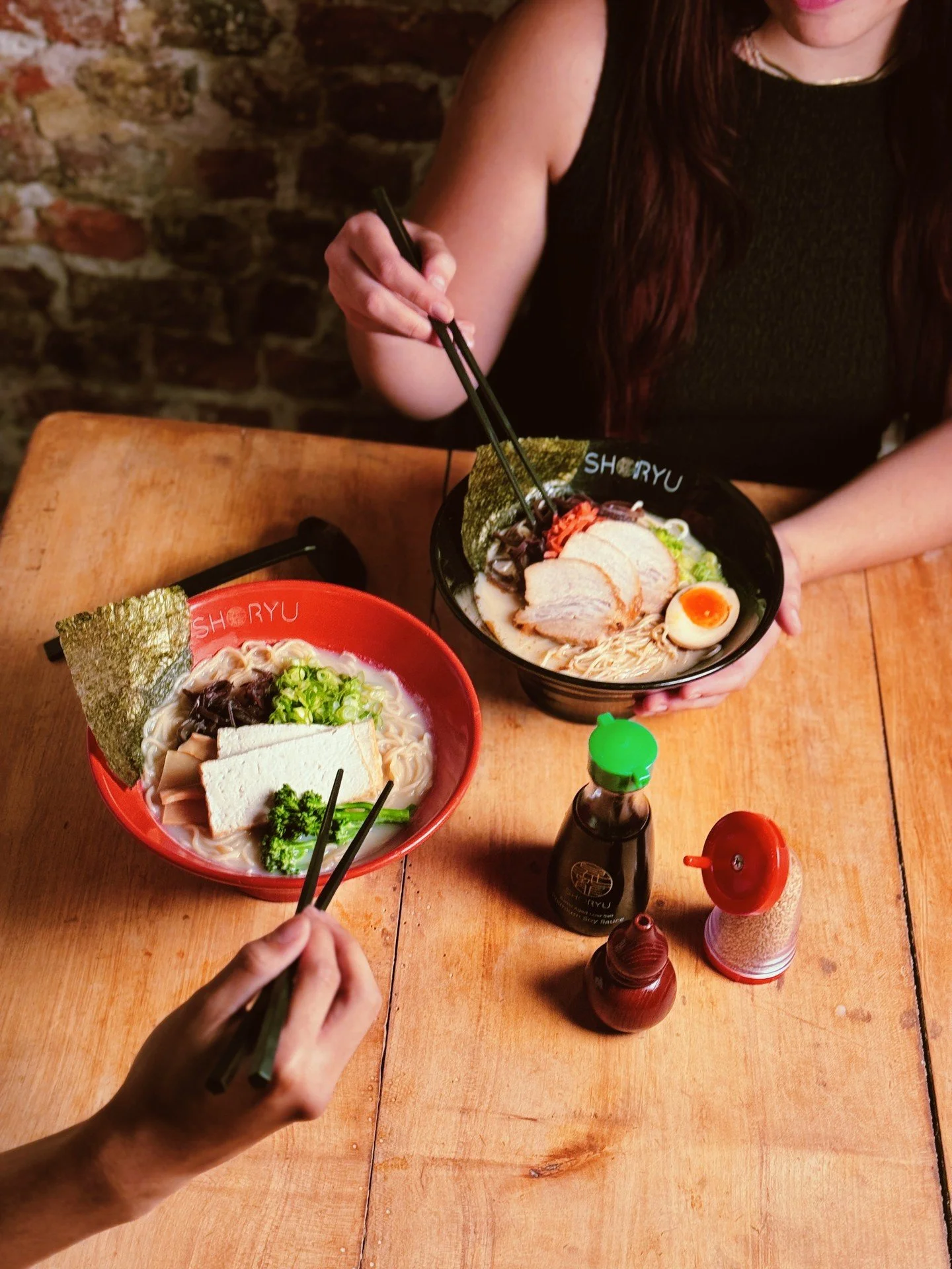 Two bowls of ramen on a wooden table with condiments, a person holding chopsticks, and a brick wall in the background.