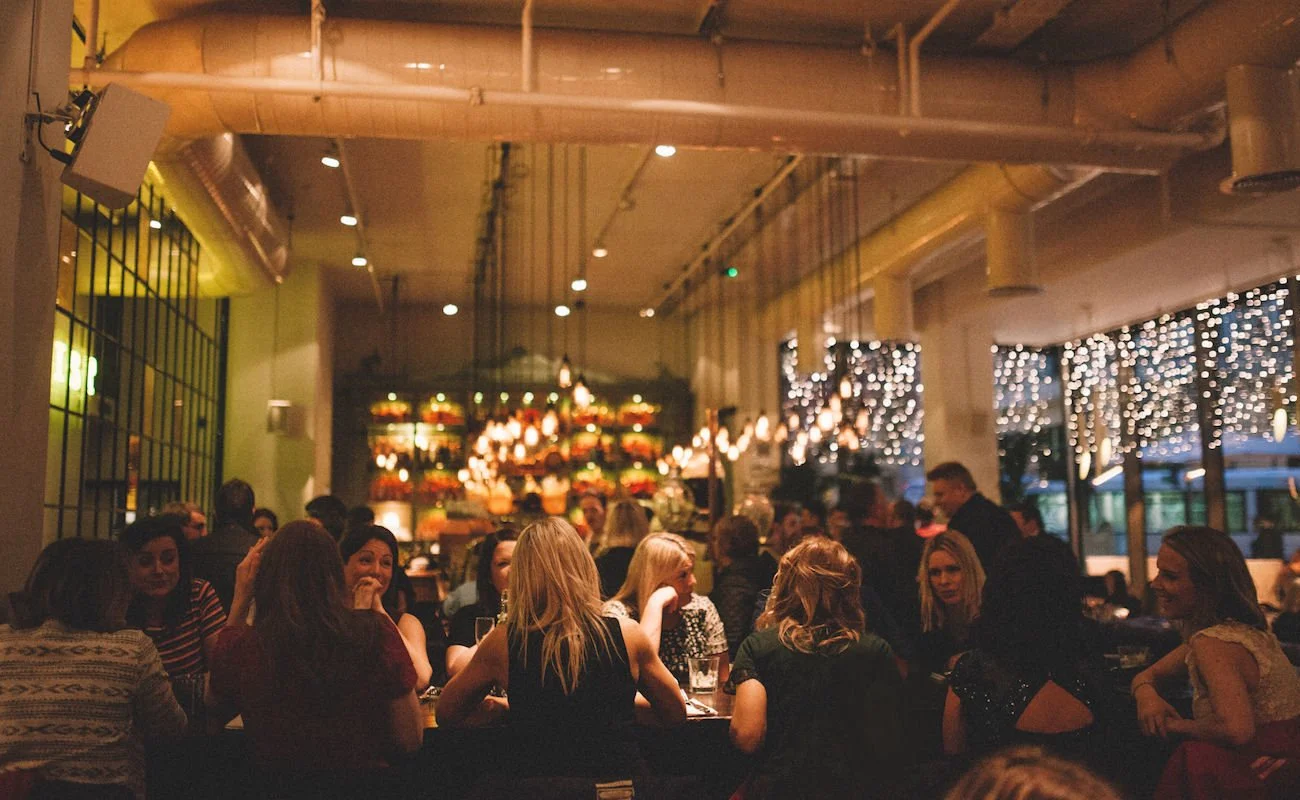 People sitting around a table in a lively restaurant or bar with string lights and shelves with plants and decor in the background.