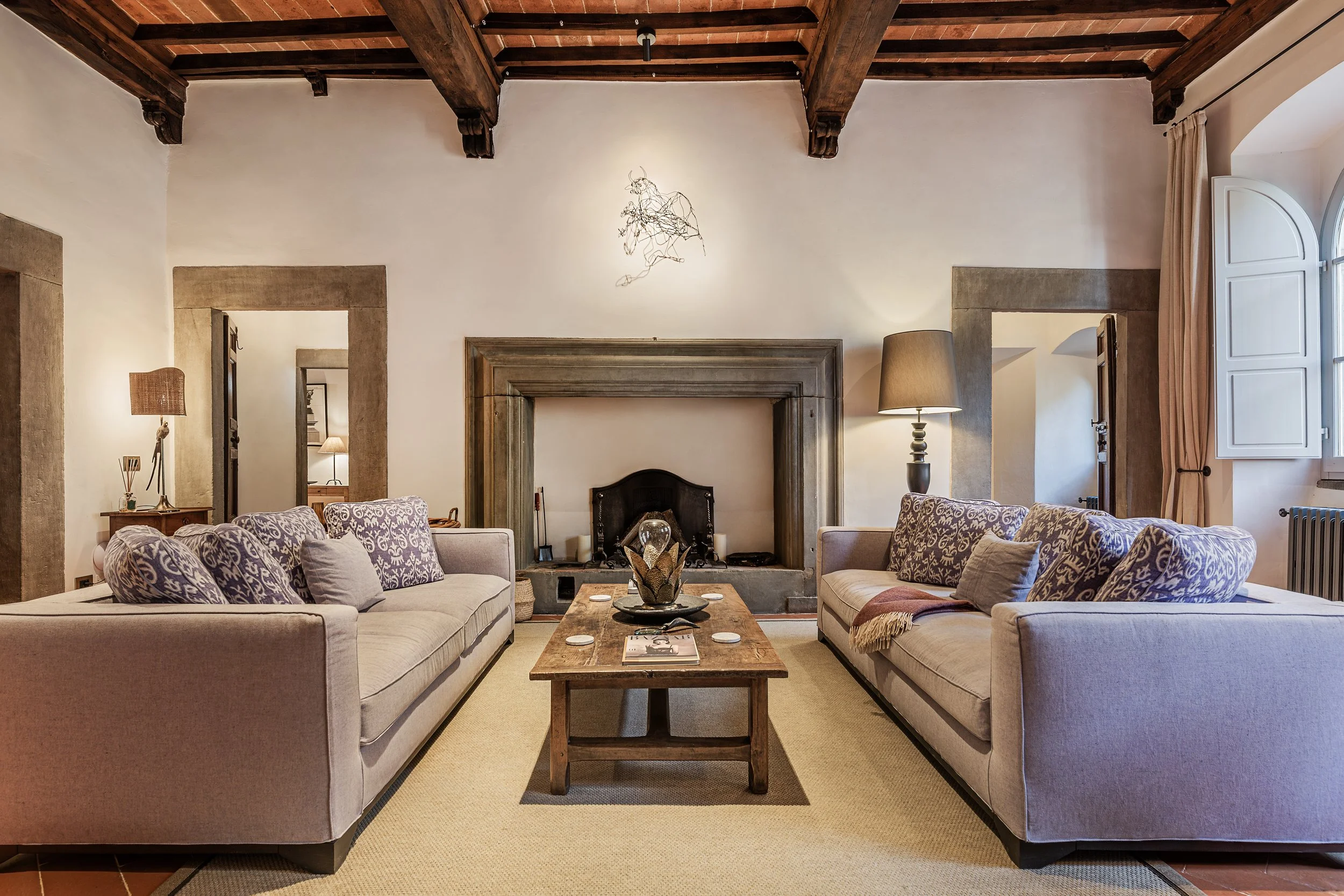Living room with two sofas with patterned cushions, a wooden coffee table, a fireplace, and lamps, with exposed wooden beams and a window with cream curtains.