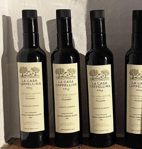 Four bottles of La Casa Cappellina extra virgin olive oil with black caps, arranged in a row on a wooden surface.