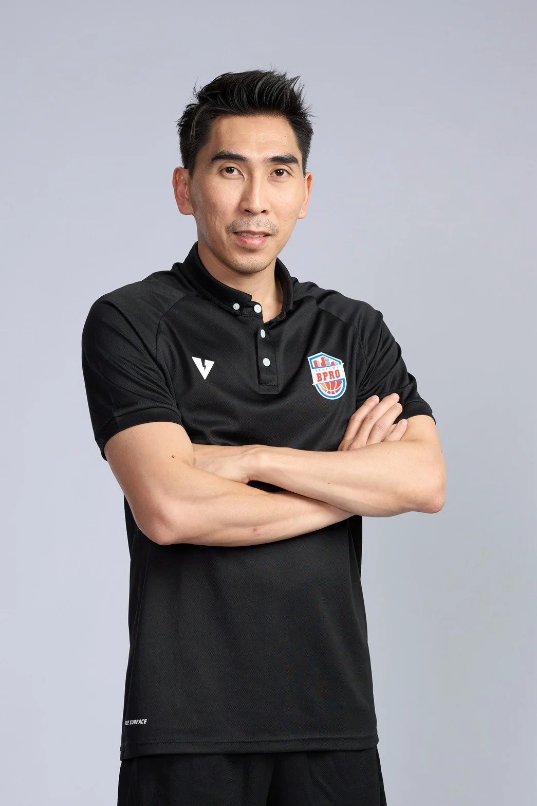 A man standing in an indoor basketball court, smiling, with a basketball hoop behind him, wearing a black sports shirt with a sports logo on the chest.