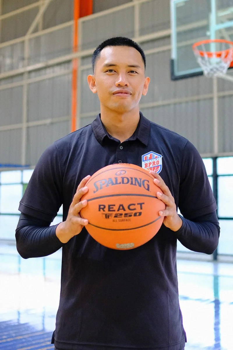 A man standing in a basketball court holding a Spalding React basketball, wearing a black shirt with a BPRO logo.