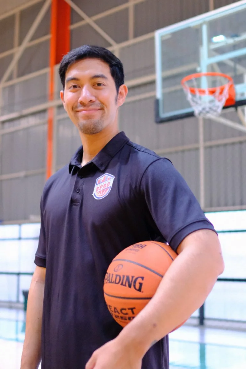 A man in a basketball court holding a basketball, smiling at the camera, wearing a black polo shirt with a US pro basketball logo.