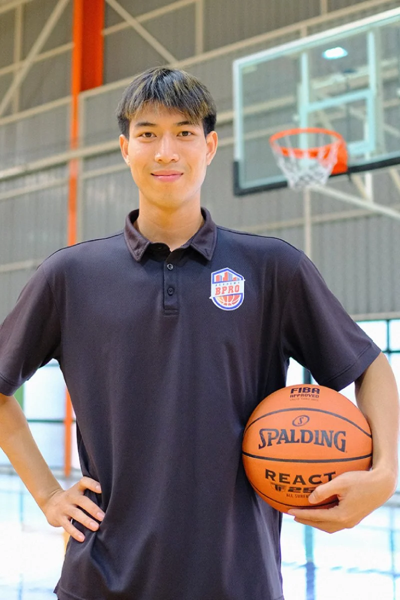 A man standing in a basketball gymnasium holding a basketball with his right hand. He is wearing a black polo shirt with a BPRO logo on it, and his left hand is resting on his hip. The background shows a basketball hoop and a court.