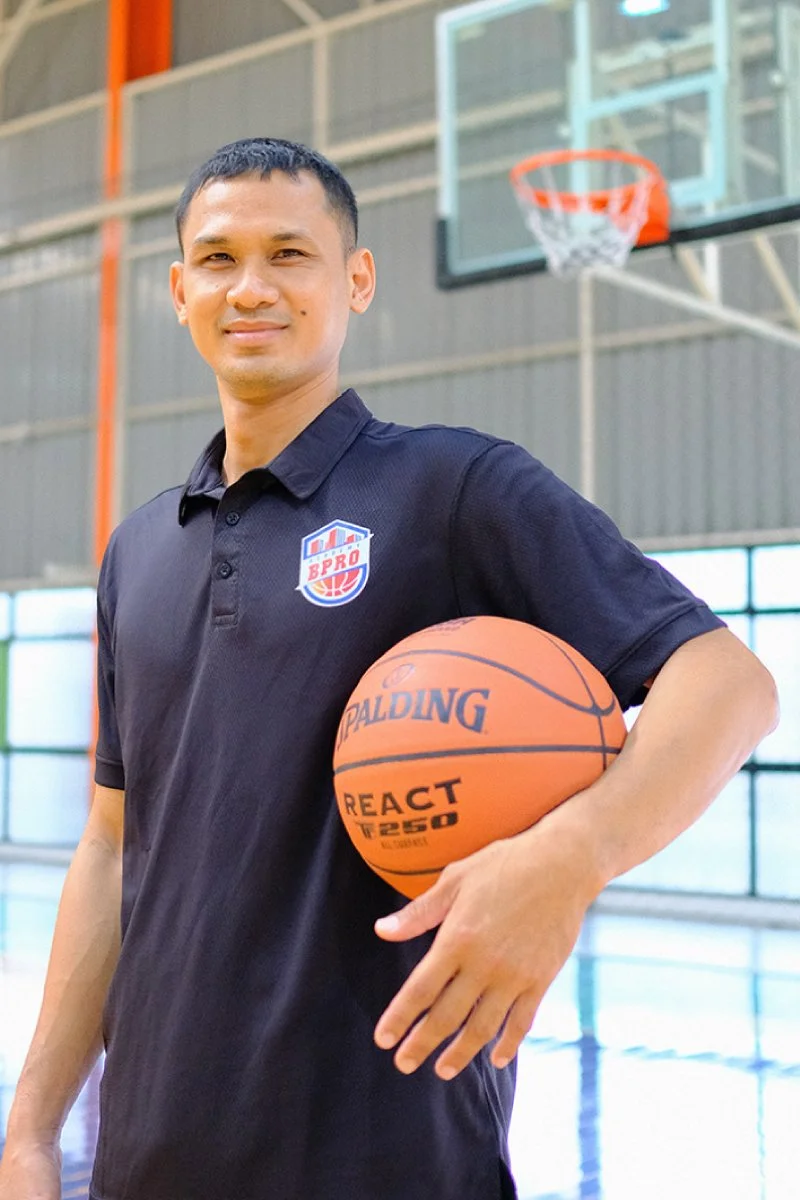 A man in a black polo shirt holding a basketball in a gymnasium with a basketball hoop in the background.