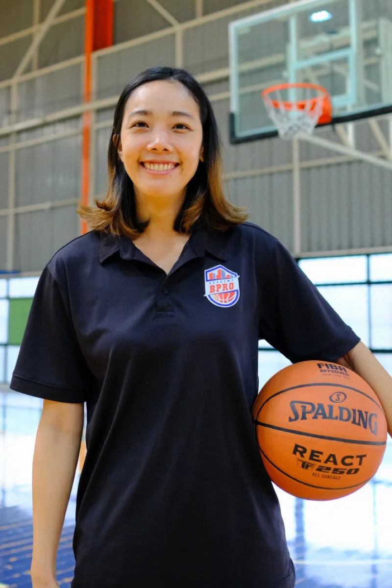 A young woman in a black polo shirt holding a basketball inside a basketball court, smiling at the camera.