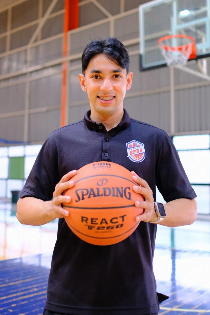 A young man smiling and holding a Spalding basketball in a gymnasium with basketball hoops and a high ceiling.