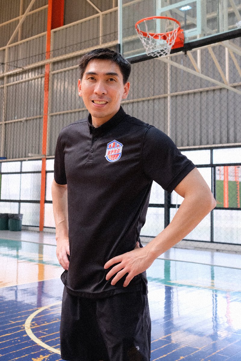 A man standing in an indoor basketball court, smiling, with a basketball hoop behind him, wearing a black sports shirt with a sports logo on the chest.