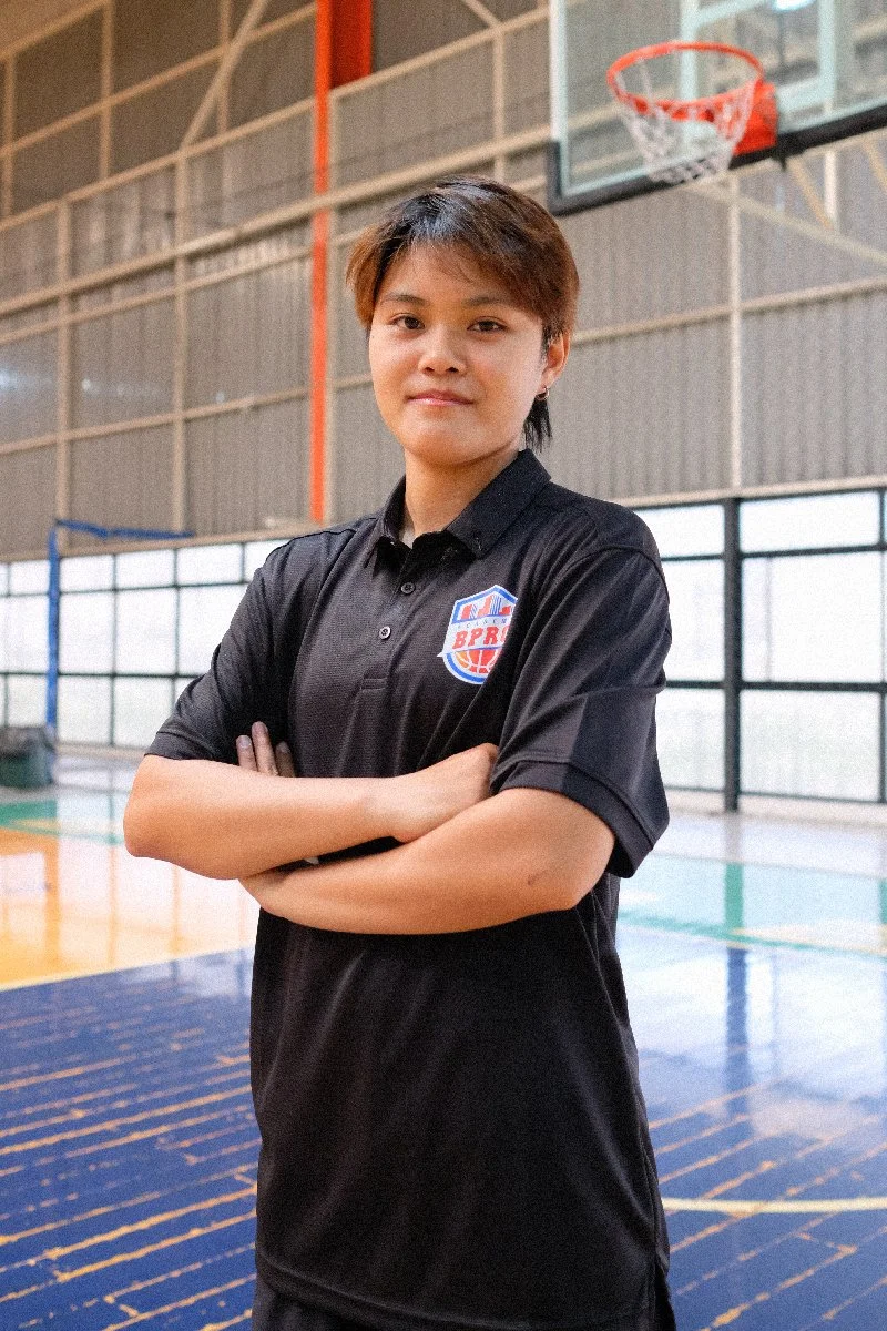Young woman standing with arms crossed in a gymnasium next to a basketball hoop.