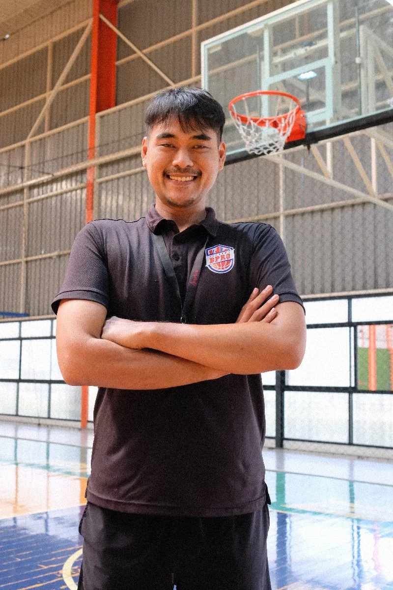 A smiling man with crossed arms standing inside a basketball gymnasium. He is wearing a black sports polo shirt with a logo that says 'BPRO' on it. In the background, there is a basketball hoop and part of a basketball court.
